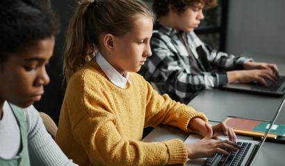 Three children of different ethnicities sit at a long desk and focus doing their schoolwork on laptop computers.