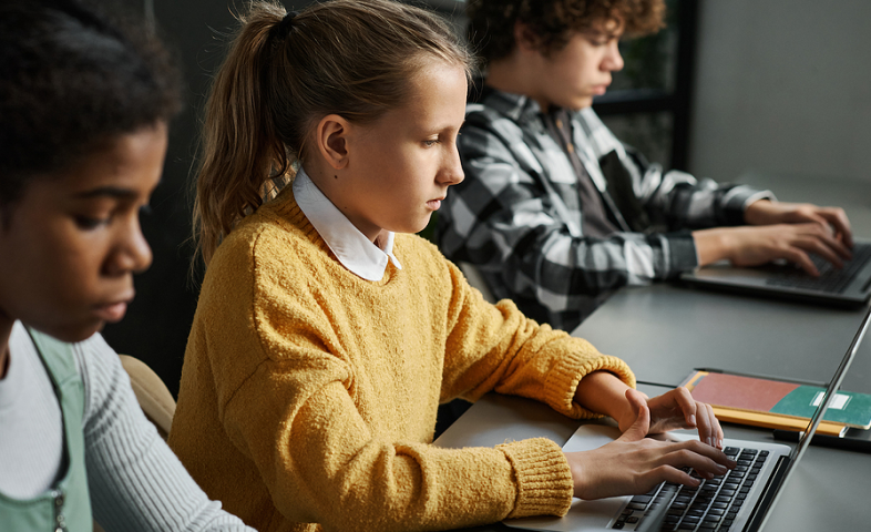 Three children of different ethnicities sit at a long desk and focus doing their schoolwork on laptop computers.