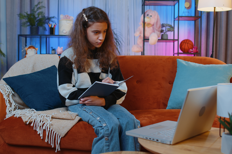 A preteen girl sits in her colorful bedroom studying and looking at her laptop screen.
