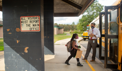 A Black female elementary school student walks toward her school bus. A school administrator stands next to the bus, watching as she boards the bus.