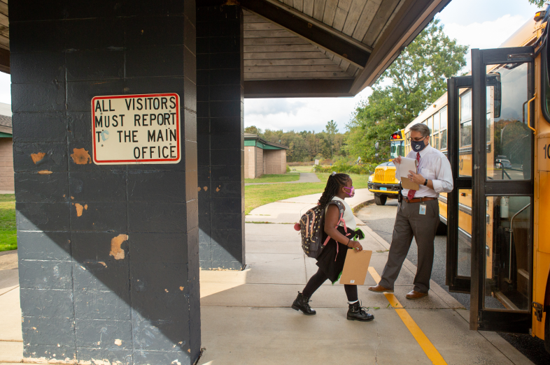 A Black female elementary school student walks toward her school bus. A school administrator stands next to the bus, watching as she boards the bus.