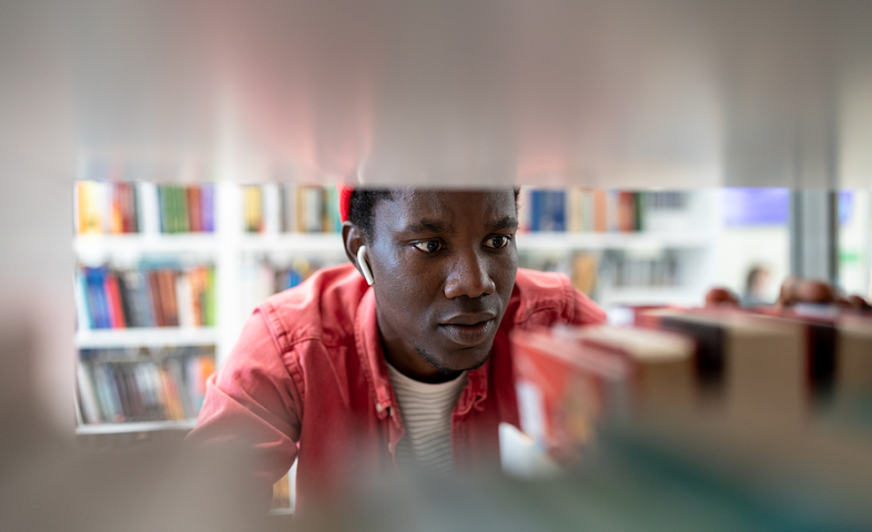 A Black college student looks for a book shelved in a campus library. He is surrounded by book stacks.
