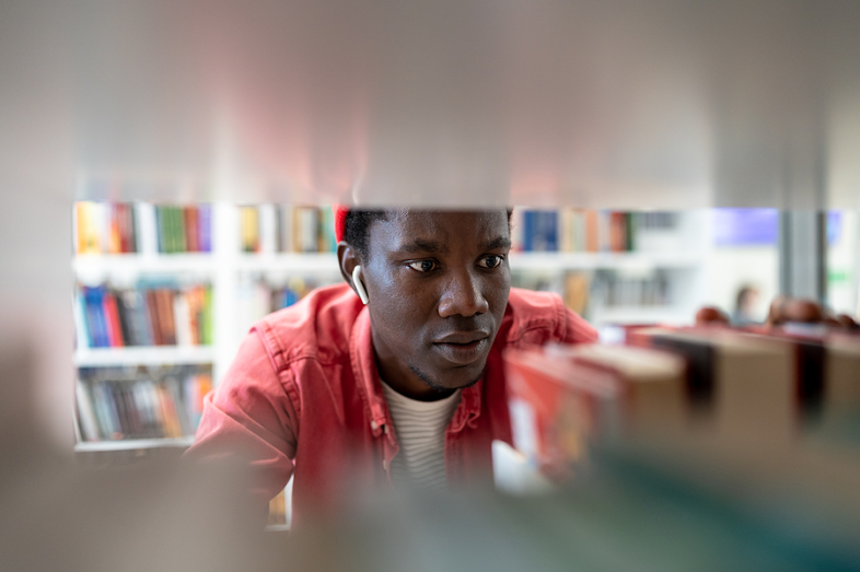 A Black college student looks for a book shelved in a campus library. He is surrounded by book stacks.