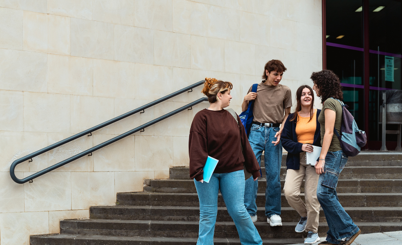 A group of college students walk down stairs and speak with each other outside their school.