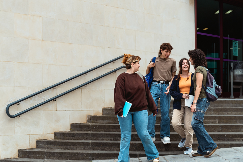A group of college students walk down stairs and speak with each other outside their school.