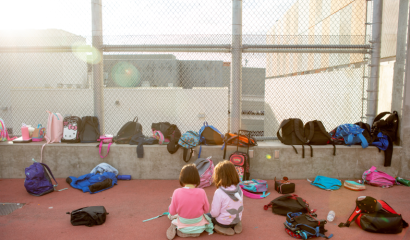 Two small elementary-aged children are outside their school, surrounded by the backpacks of their classmates.