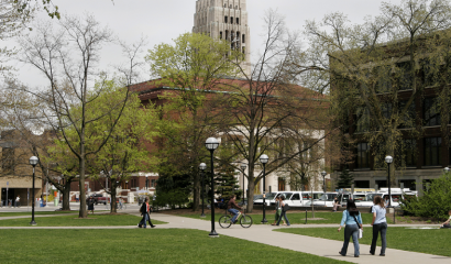 Gray skies appear over the University of Michigan campus as few walk along paved paths. They're surrounded by brick buildings and trees with sparse leaves.
