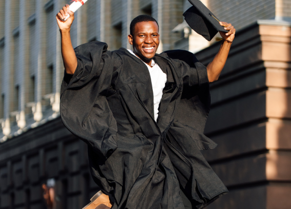 A Black young man jumps with joy as he wears his black graduation gown and holds a diploma and cap