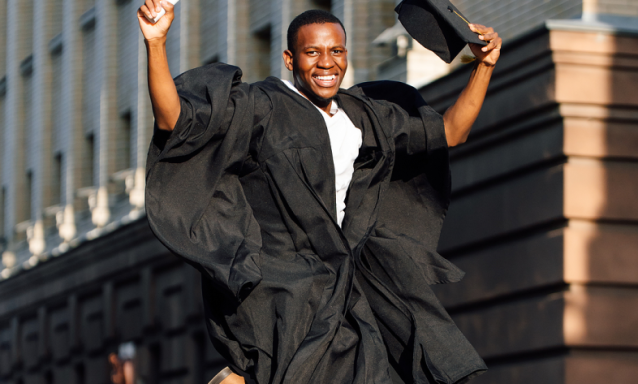 A Black young man jumps with joy as he wears his black graduation gown and holds a diploma and cap