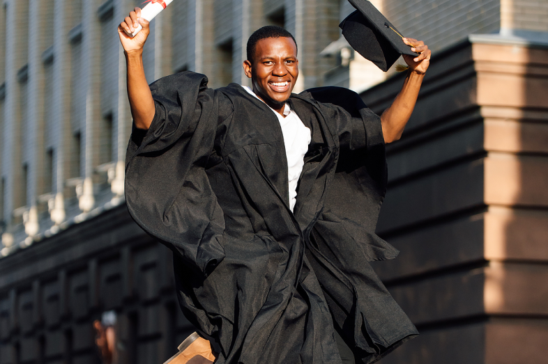 A Black young man jumps with joy as he wears his black graduation gown and holds a diploma and cap