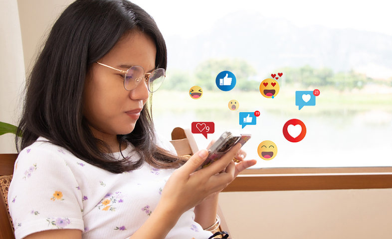 A young woman looks down at her cellphone. Social media graphics create a cloud of around her.