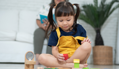 A little toddler girl, who is surrounded by wooden, colorful blocks, picks up a red block, staring at it.