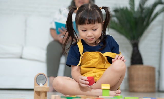 A little toddler girl, who is surrounded by wooden, colorful blocks, picks up a red block, staring at it.