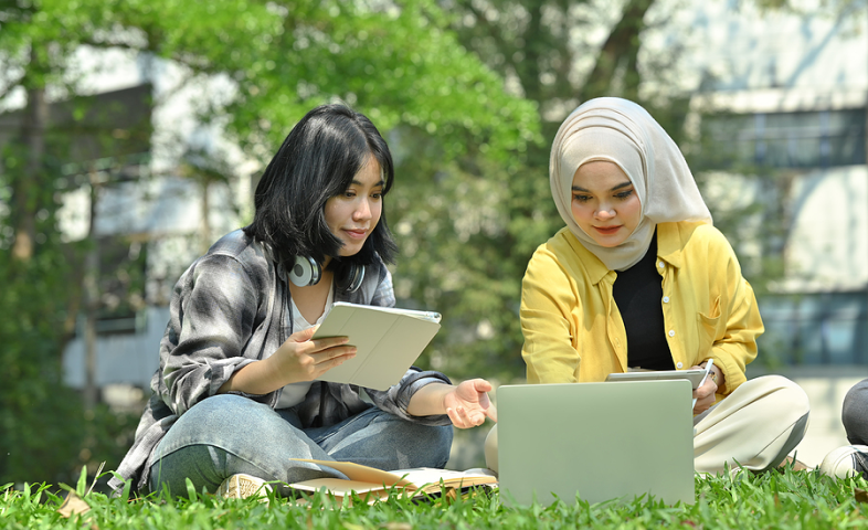 Two Asian college students sit outdoors on grass, working on an assignment