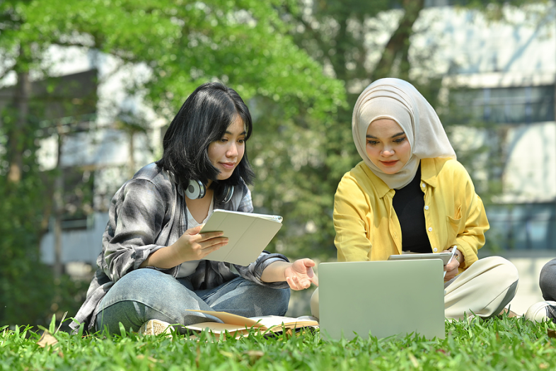 Two Asian college students sit outdoors on grass, working on an assignment