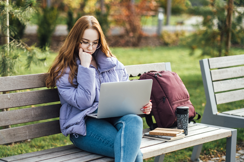 A young woman sits on a park bench, staring at her laptop screen.