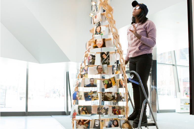 A teenage girl stands on a ladder, placing the finishing touches on wooden double-helix style sculpture that's taller than a Christmas tree.