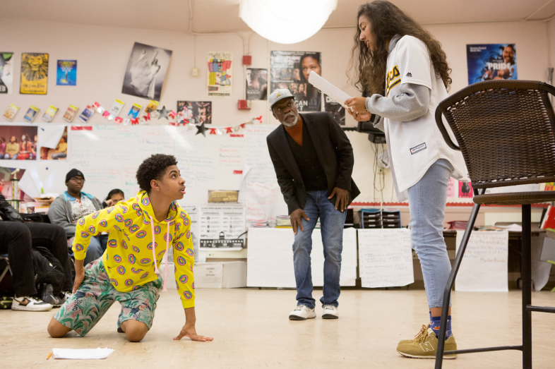 Students act out a scene from a play during rehearsal. One kneels on the floor while another stands reading a script. Their teacher looks on in the background.