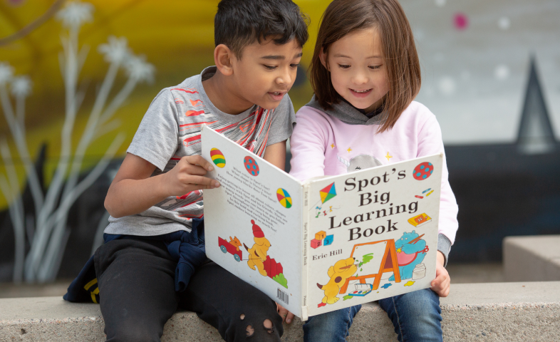A elementary school aged boy and girl sit outside, staring at a large book.