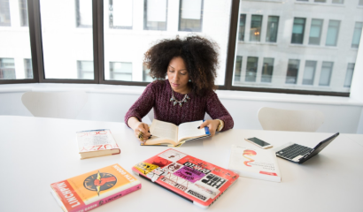 A Black woman holds a book open. She is surrounded by more books and an open laptop.