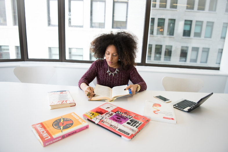 A Black woman holds a book open. She is surrounded by more books and an open laptop.