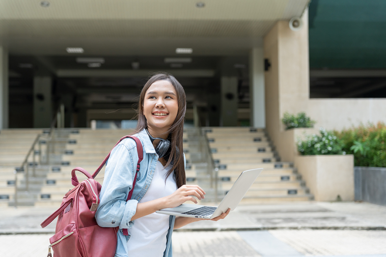 A college student holds a laptop as she stands in front of a staircase on what appears to be a college campus.
