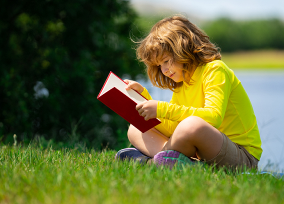 A small boy sits on green grass reading a book. A body of water is behind him.