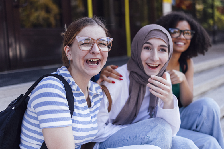Three multiracial students sit laughing on outdoor steps.