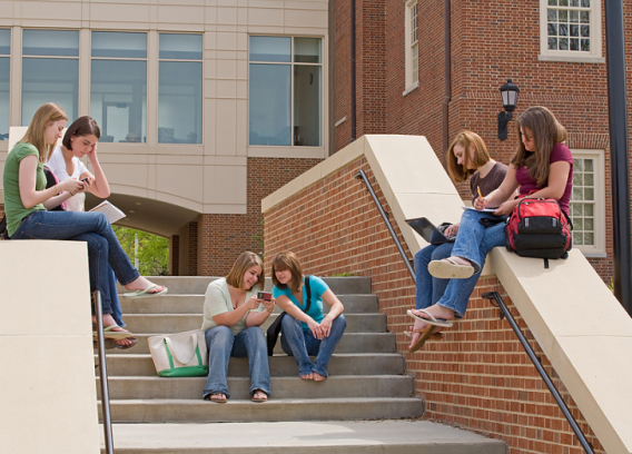 A group of college girls sit on the steps of a campus building.