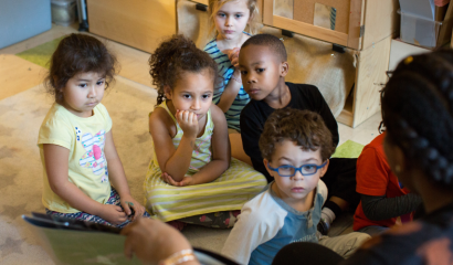 Children sit on the floor, listening to their teacher read.