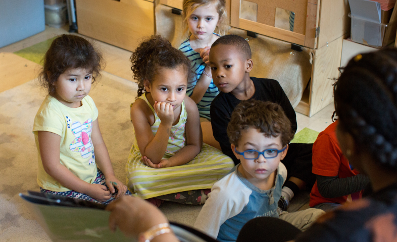 Children sit on the floor, listening to their teacher read.