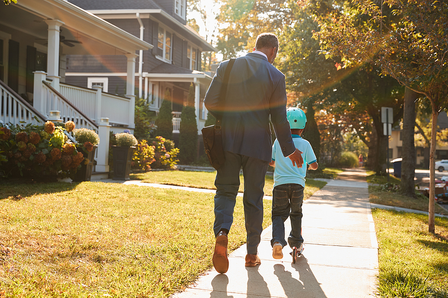 Father walks his son to school along a quiet suburban street filled with green lawns and trees.
