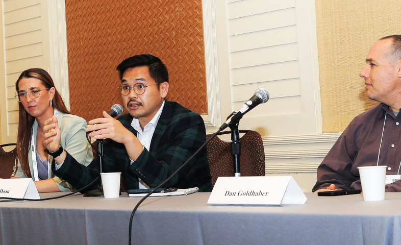 Three people sit at a table while one speaks.