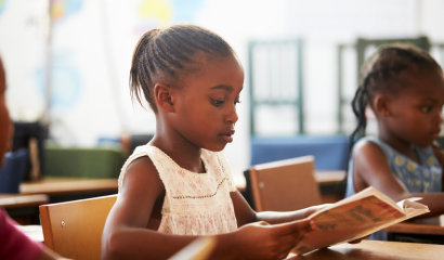 Two Black elementary-aged girls sit at desks and read.