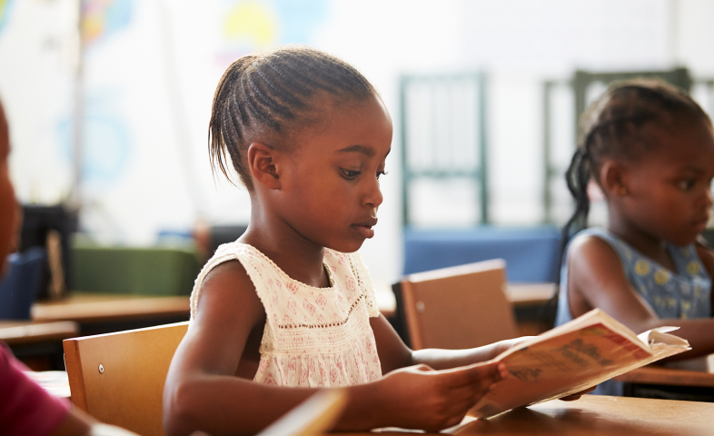 Two Black elementary-aged girls sit at desks and read.