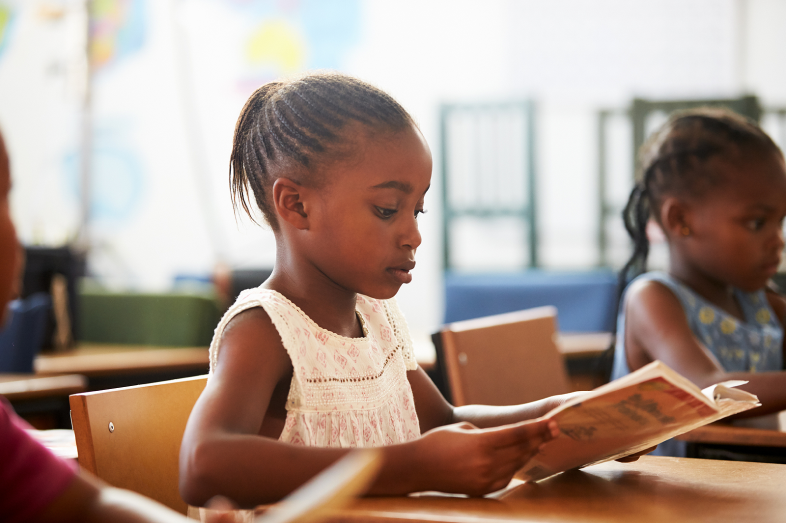 Two Black elementary-aged girls sit at desks and read.