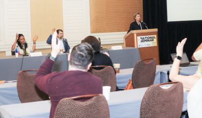 Individuals sitting at rows of tables raise their hand as a speaker talks to them.