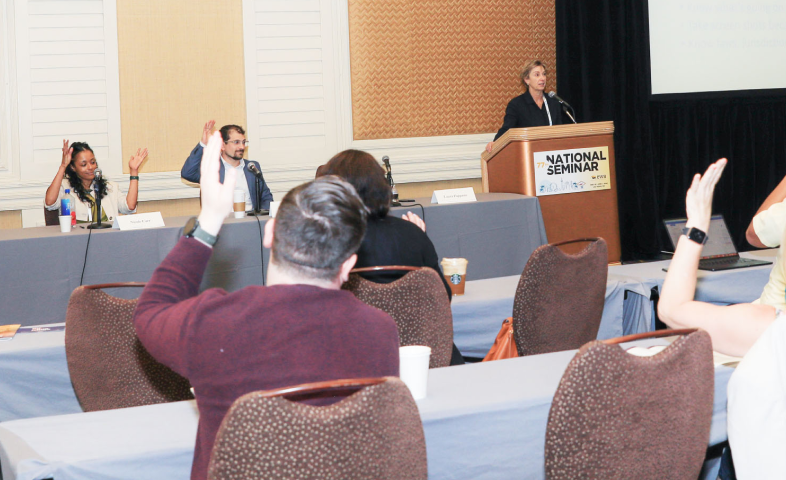 Individuals sitting at rows of tables raise their hand as a speaker talks to them.