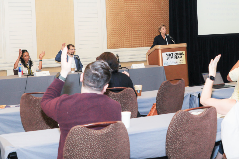 Individuals sitting at rows of tables raise their hand as a speaker talks to them.