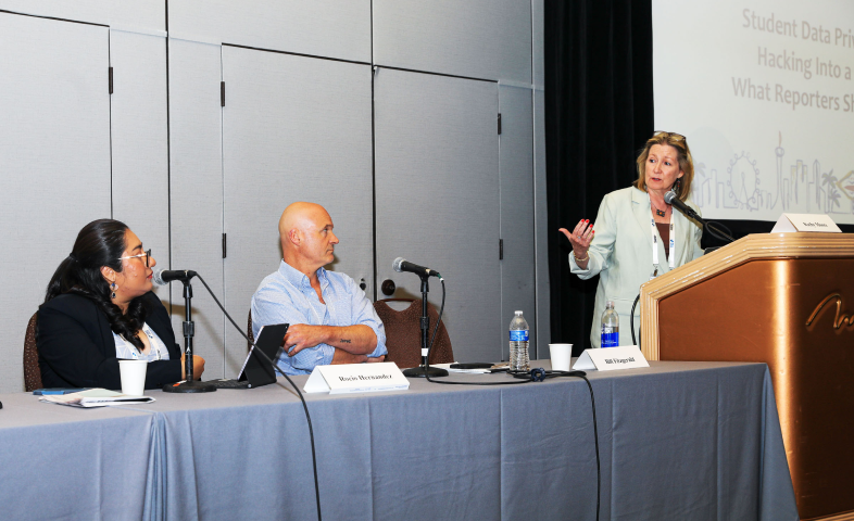 Two people sit at a table while another stands at a podium.