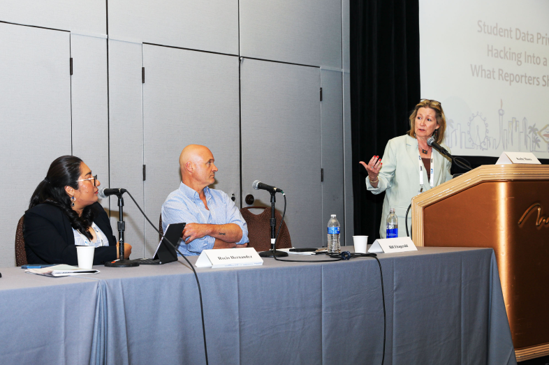 Two people sit at a table while another stands at a podium.