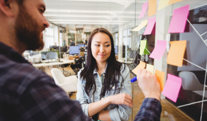 A man writes on a sticky note as a woman looks on.