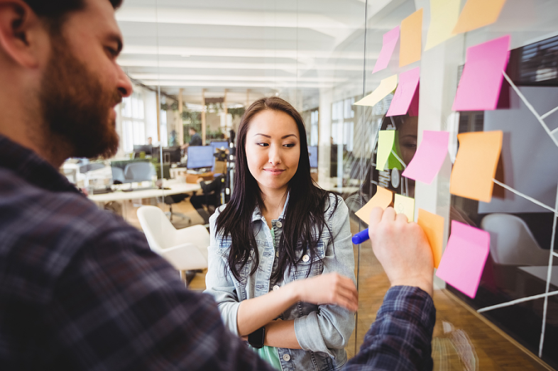 A man writes on a sticky note as a woman looks on.