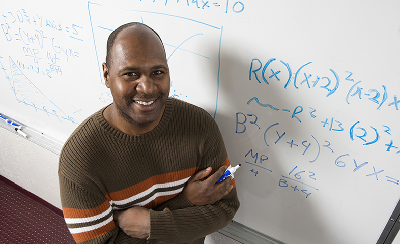 A Black male teacher stands in front of a whiteboard holding a marker after writing math equations.