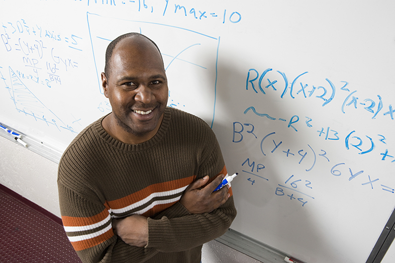 A Black male teacher stands in front of a whiteboard holding a marker after writing math equations.