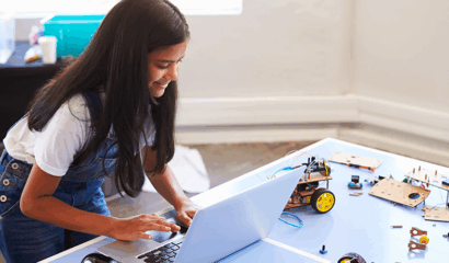A girl hunches over her computer, near robot parts.