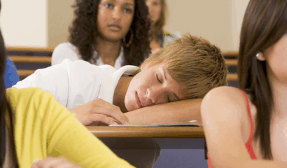 A teen boy sleeps on his desk at school.