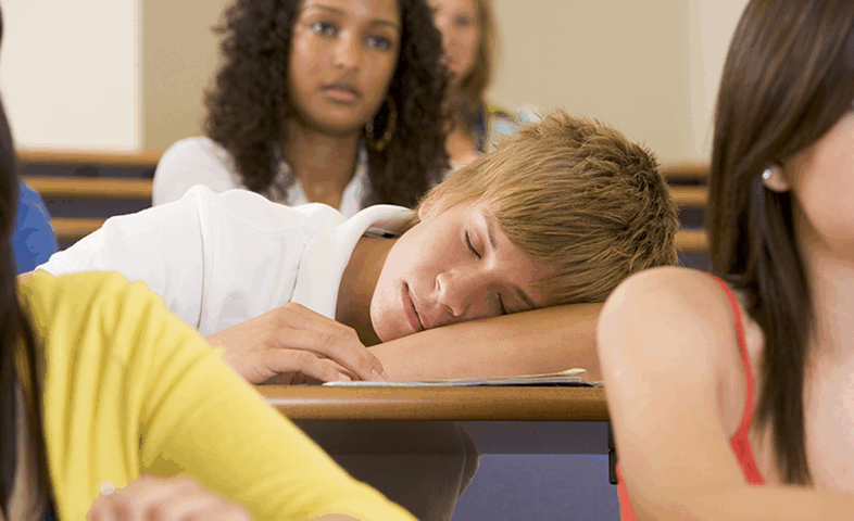 A teen boy sleeps on his desk at school.