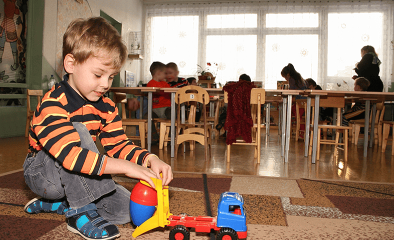 A little boy plays with a toy car as a teacher and two students are near school desks in the background.