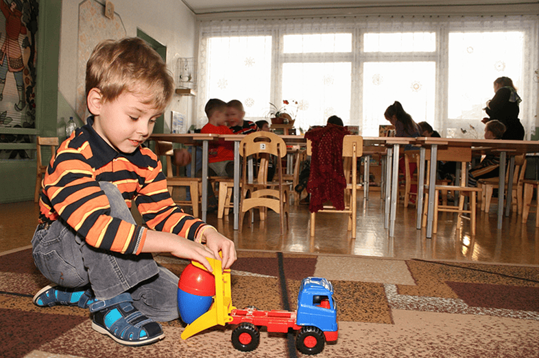 A little boy plays with a toy car as a teacher and two students are near school desks in the background.